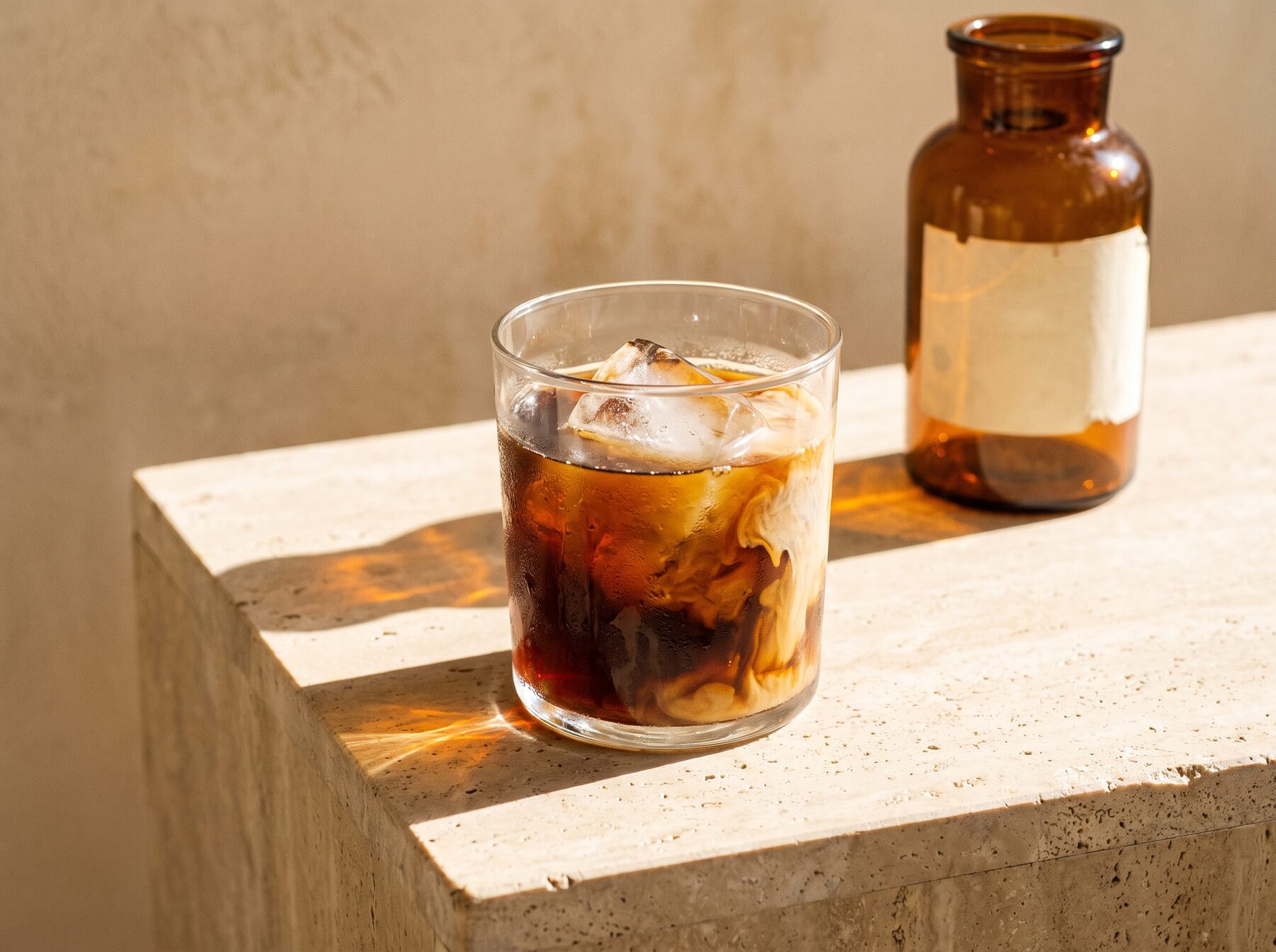 A glass of cold brew over ice with cream swirling through it, sunlight throwing amber light on stone