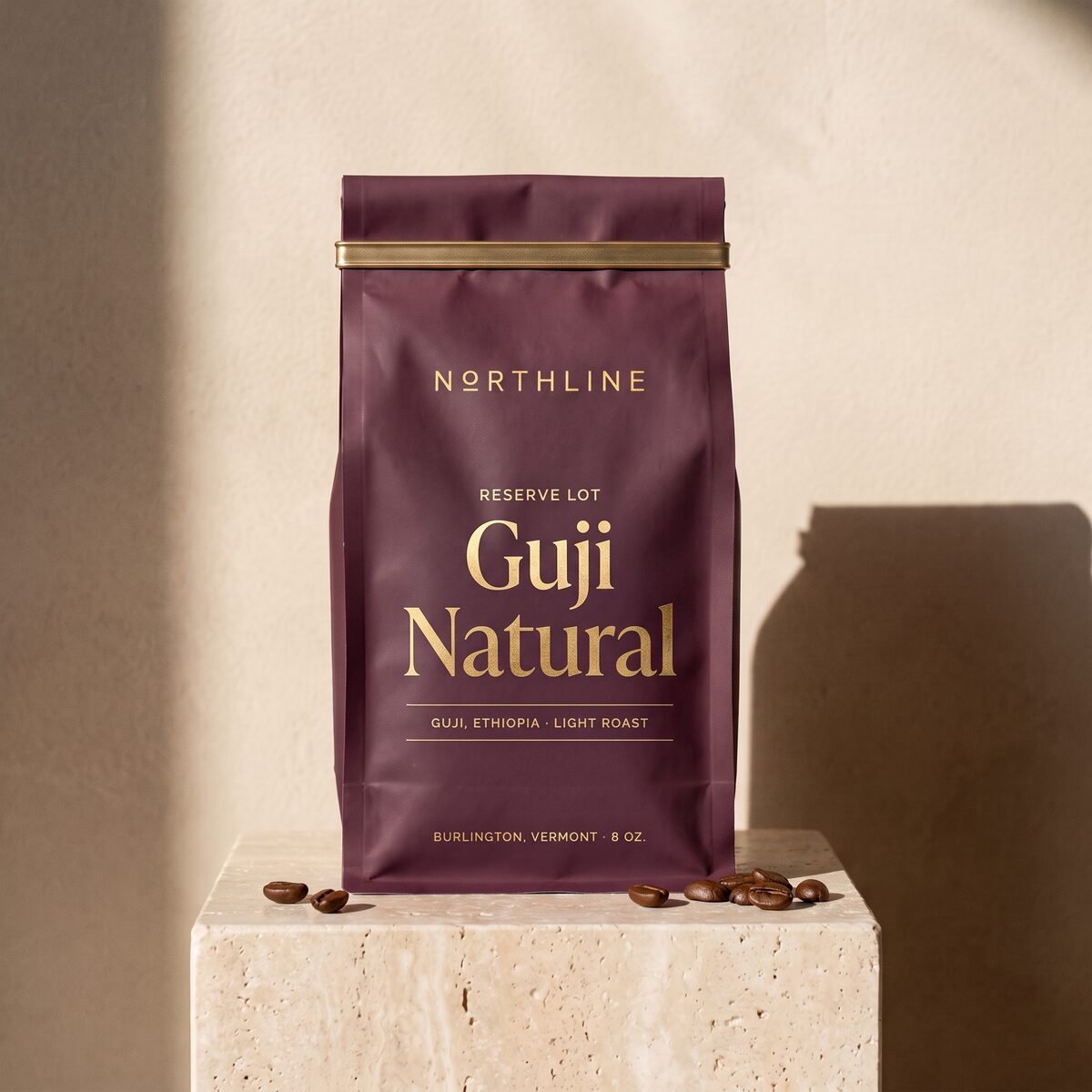 Guji Natural, marked Reserve Lot in gold, in a matte plum Northline coffee bag with a brass tie, on a travertine block against a plaster wall