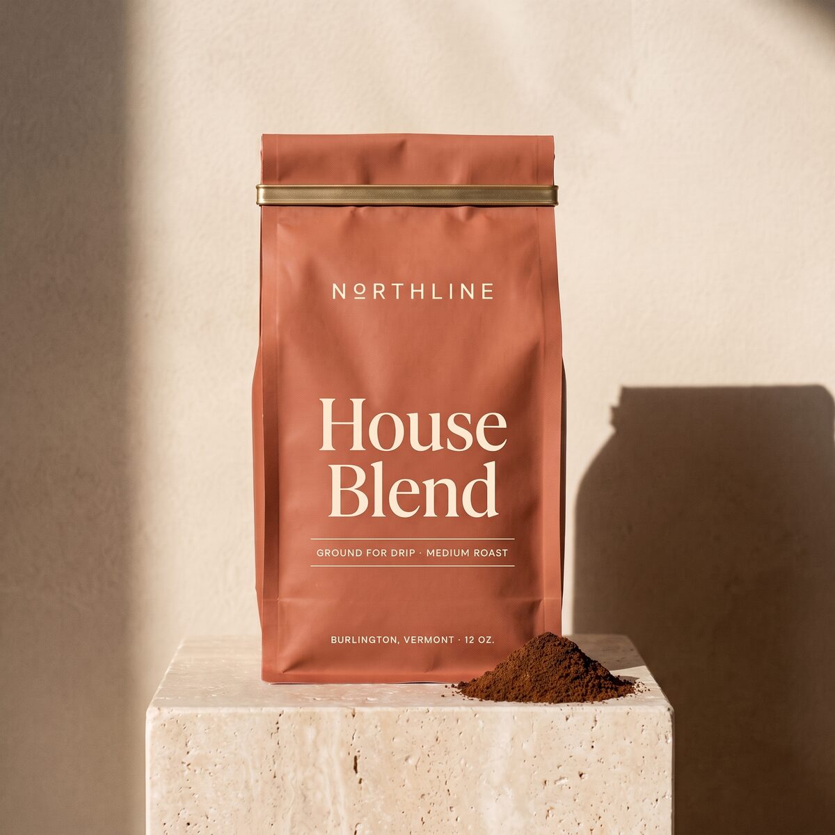 House Blend, ground for drip, in a matte terracotta Northline coffee bag with a brass tie, on a travertine block against a plaster wall, with a small mound of ground coffee