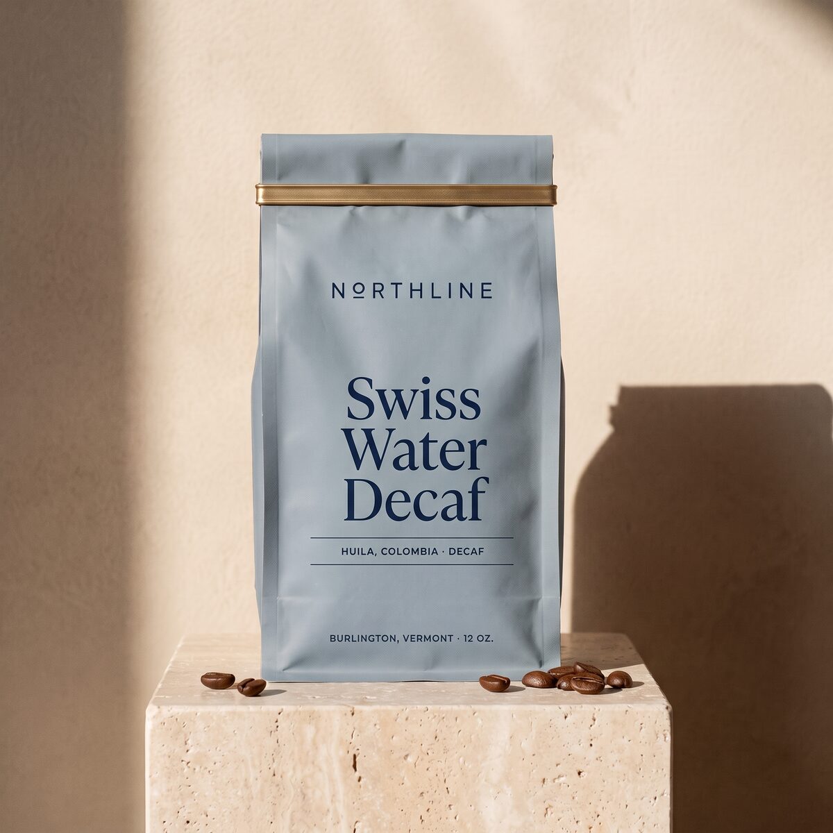 Swiss Water Decaf in a matte blue-grey Northline coffee bag with a brass tie, on a travertine block against a plaster wall